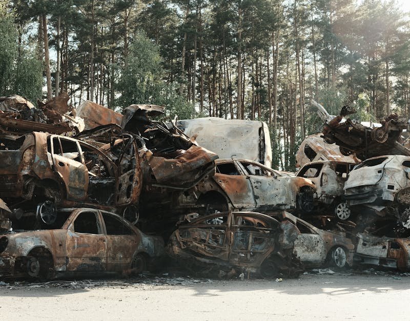 A pile of rusted, destroyed cars abandoned in a forest junkyard, symbolizing waste and decay.