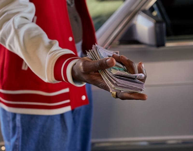 Close-up of a person offering a stack of cash in front of a car, symbolizes financial transaction.