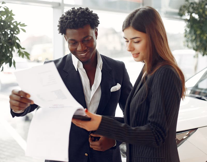 Cheerful smiling multiethnic businessman in classy suit and professional elegant female consultant standing close and reading contract details in car showroom