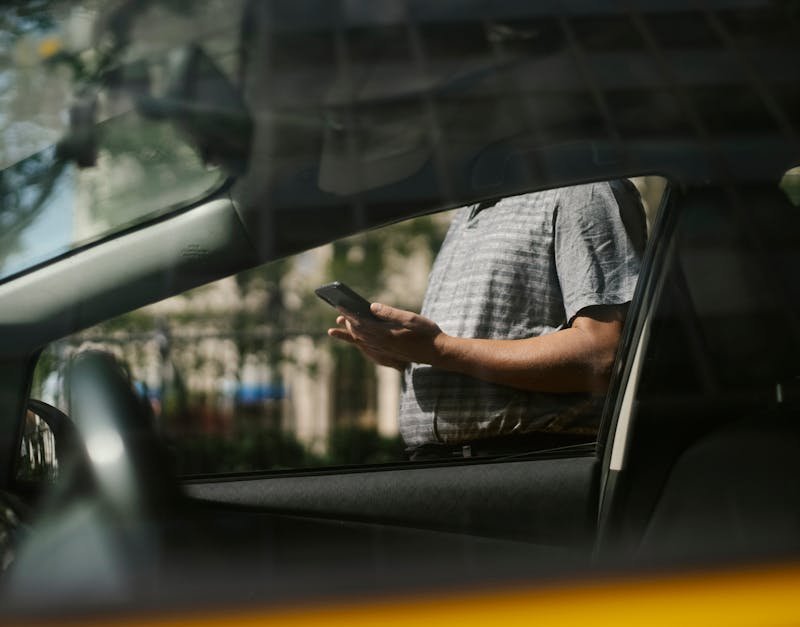 A man using a smartphone inside a car, visible through the window in an urban setting.