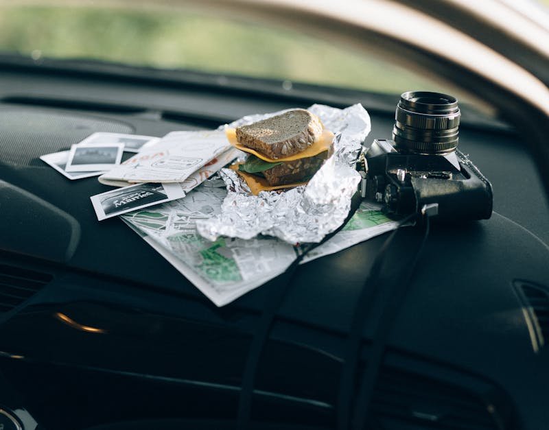 A camera and a sandwich wrapped in foil placed on a car dashboard with maps and photos.