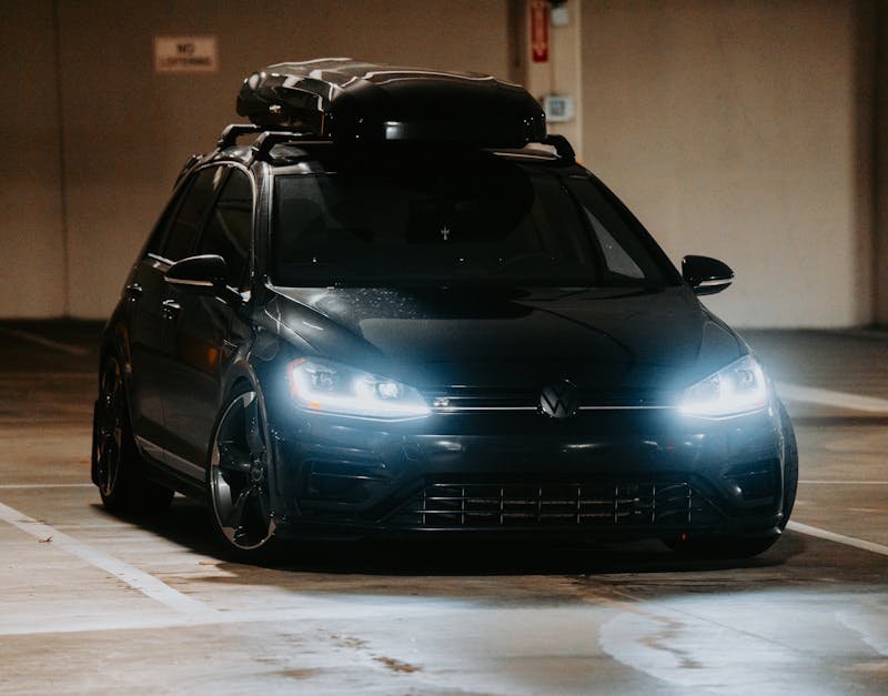 A stylish black car with headlights on, parked indoors in a dimly lit garage, exuding elegance.