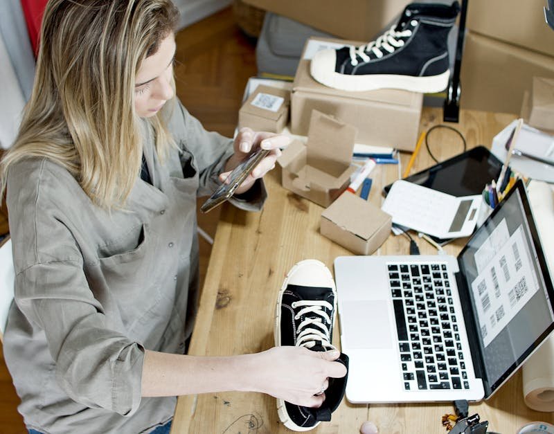Woman photographing shoes for online sale in home workspace with laptop and packages.