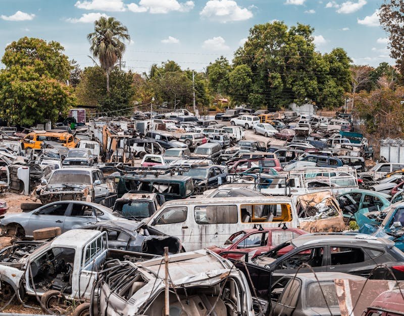 A busy junkyard filled with a variety of old cars under a clear sky, surrounded by trees.