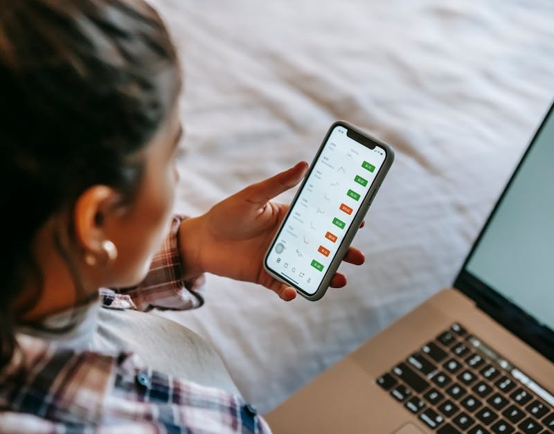 Female analyzing stock market data on her smartphone and laptop indoors. Ideal for finance, technology, and investment themes.