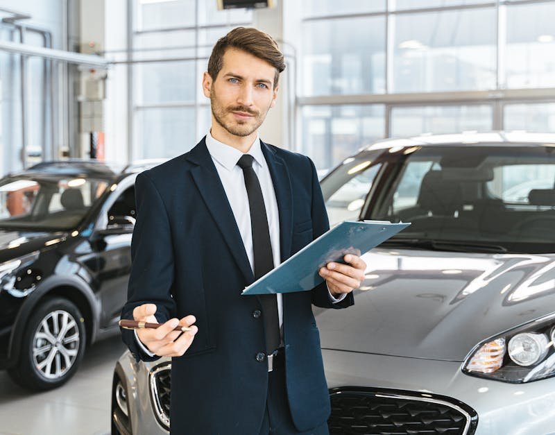 Professional car dealer in business suit holding clipboard in a bright car showroom.