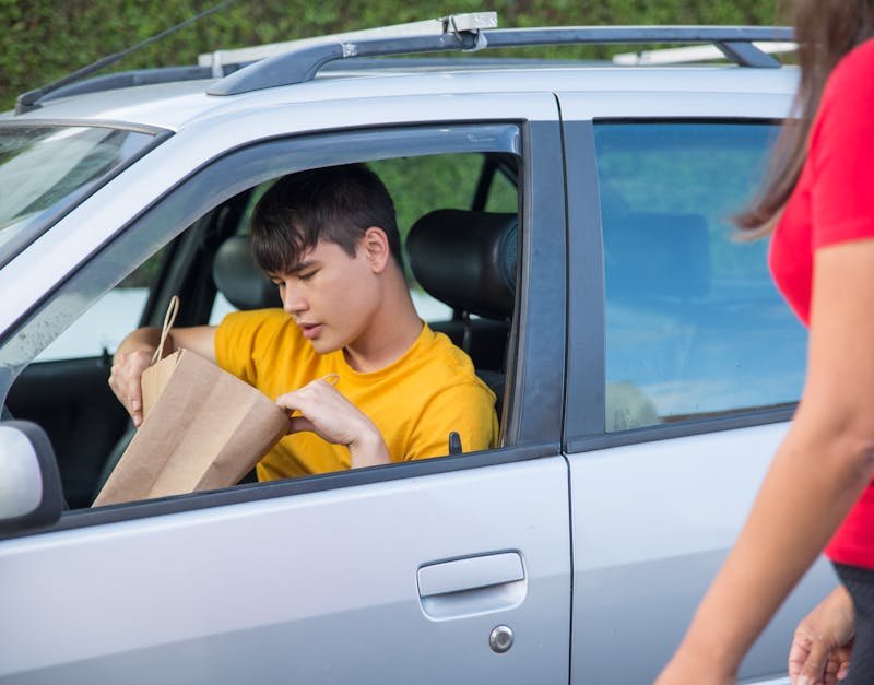 A driver in a yellow shirt delivers a paper bag order to a customer beside a car.