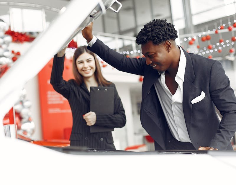 Positive smiling diverse stylish man and female dealer checking under car motor hood and smiling while choosing new car in car showroom