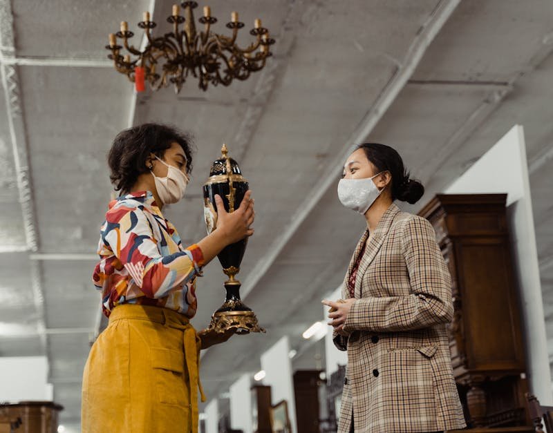 Two women in masks evaluating an antique vase in a shop, showcasing vintage fashion.