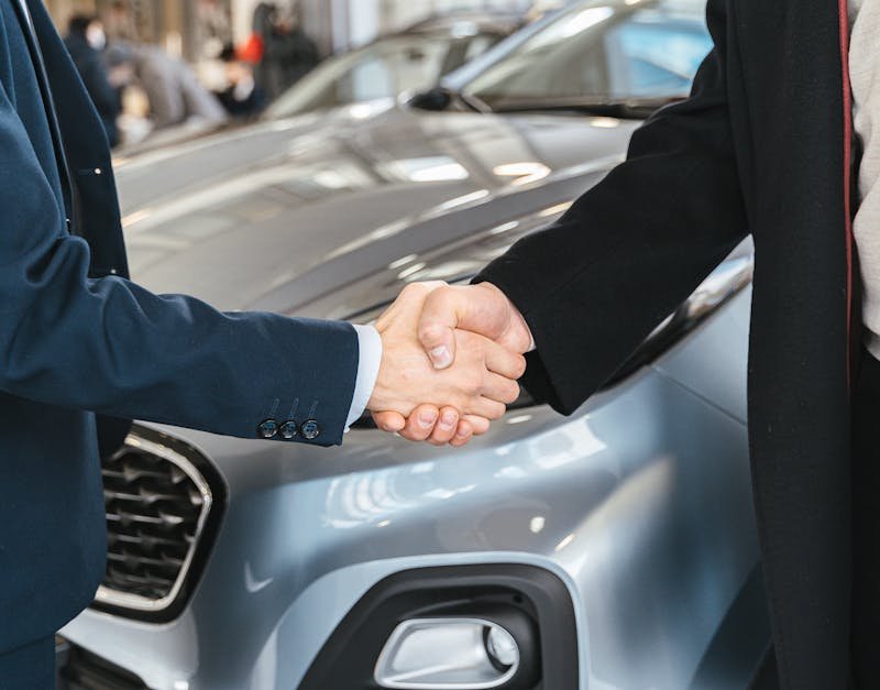 A close-up of two people shaking hands in front of a car in an indoor setting.