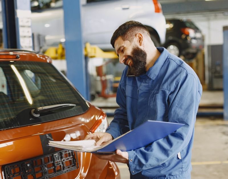 Smiling auto mechanic inspects a car in a service garage, wearing coveralls and holding documents.