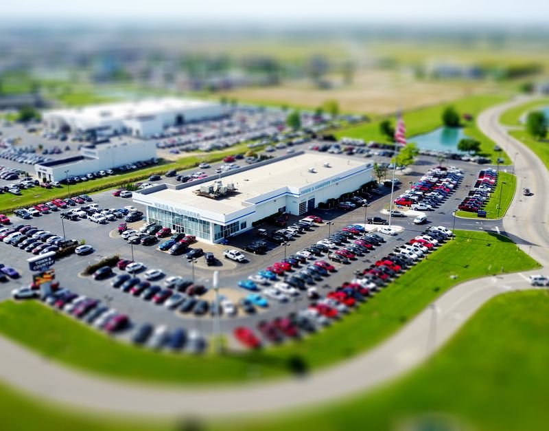 Aerial high-angle view of a bustling car dealership surrounded by parked cars in a green landscape.